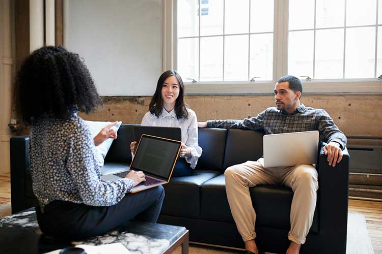 Three people on couches with laptops having discussion.