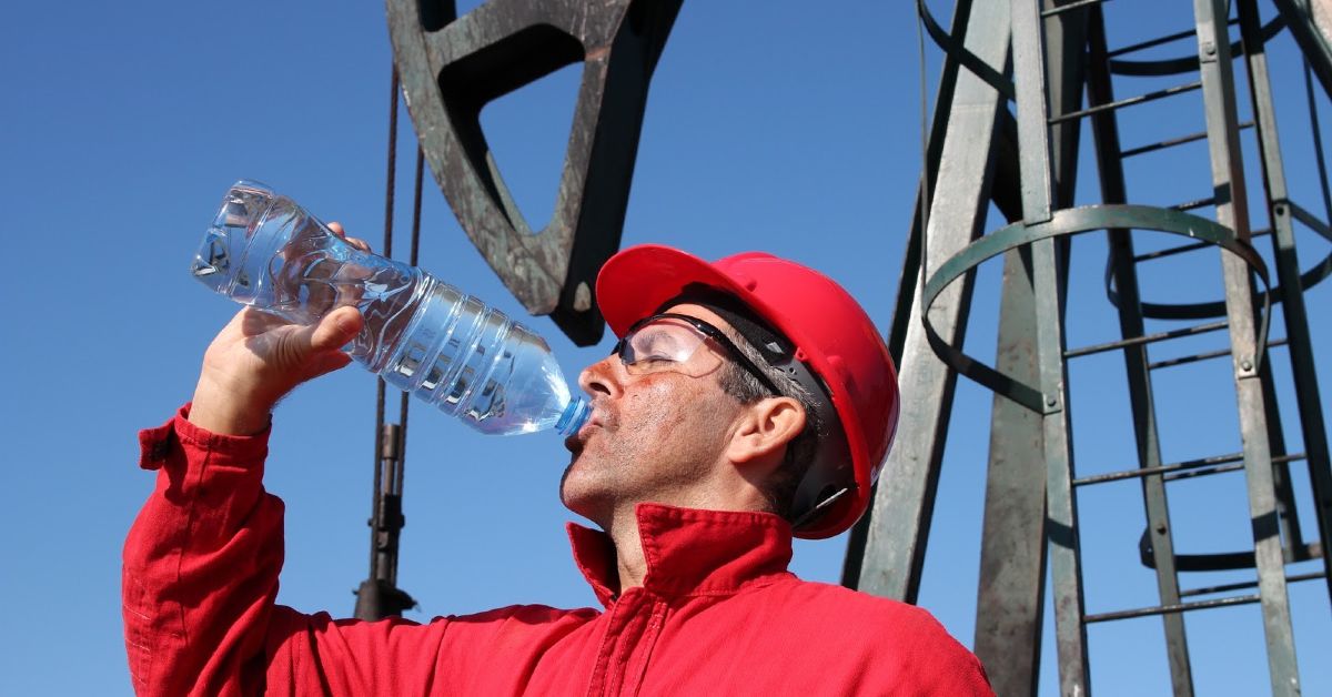 A man wearing a hard hat and glasses drinks from a bottled water.