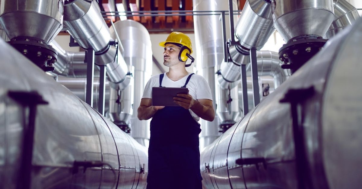 A man wearing a hard hat and protective ear muffs surveys equipment in an industrial plant.