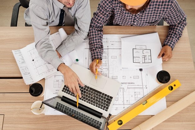 Top-down view of two people discussing printed architectural plans and information on a computer screen.