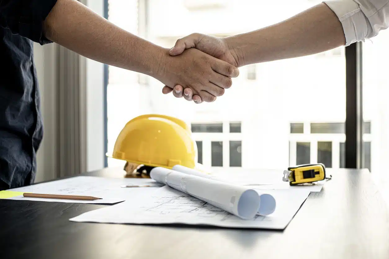 Two people shaking hands over construction blueprints and a yellow hard hat.