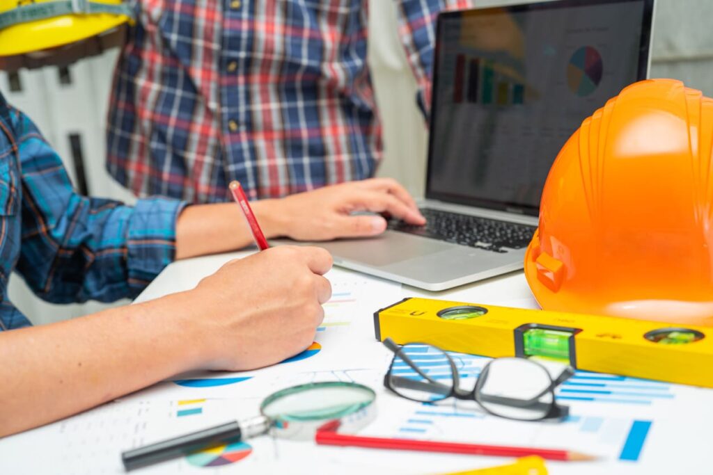 A person writing notes and using a laptop on a desk with a construction hat and level.