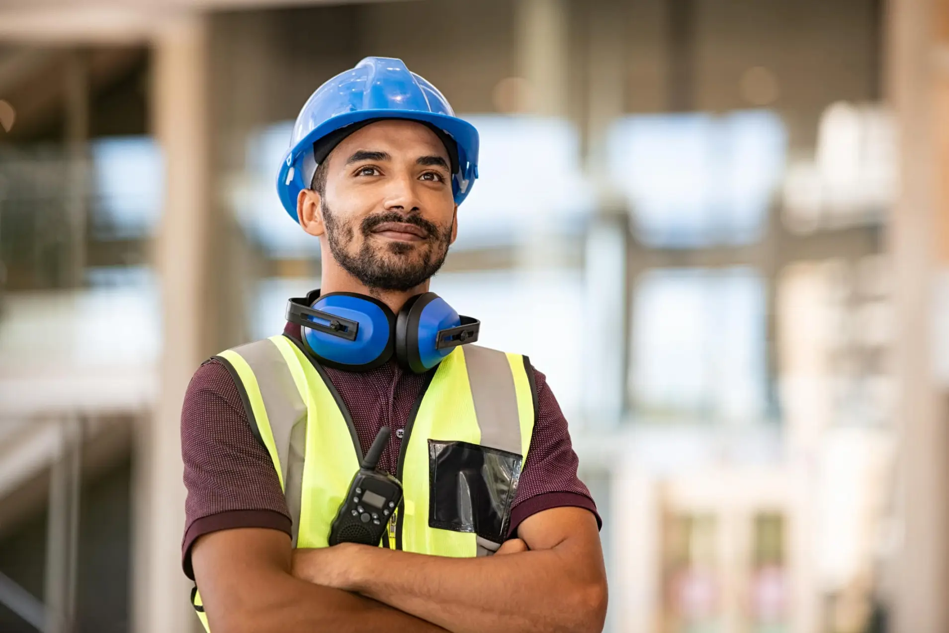 A construction site worker smiling and standing with folded arms, wearing a safety vest and helmet.