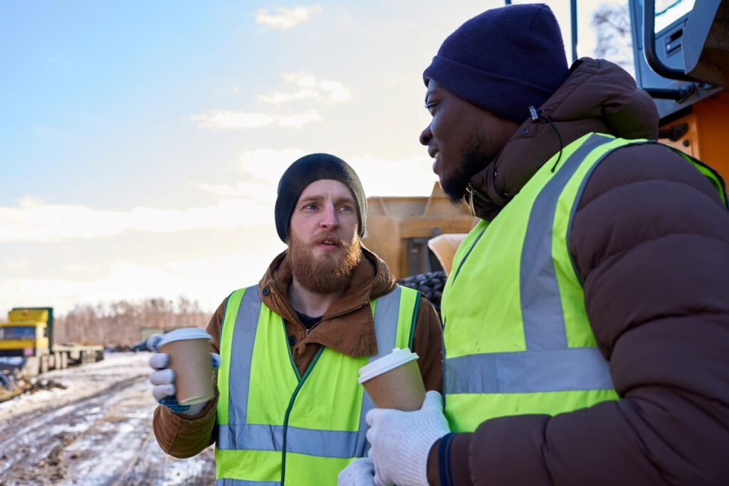 Two construction workers in winter gear, holding coffee cups, take a break at a snowy site.