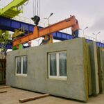 A crane lifts concrete modular wall panels with windows at a construction site.