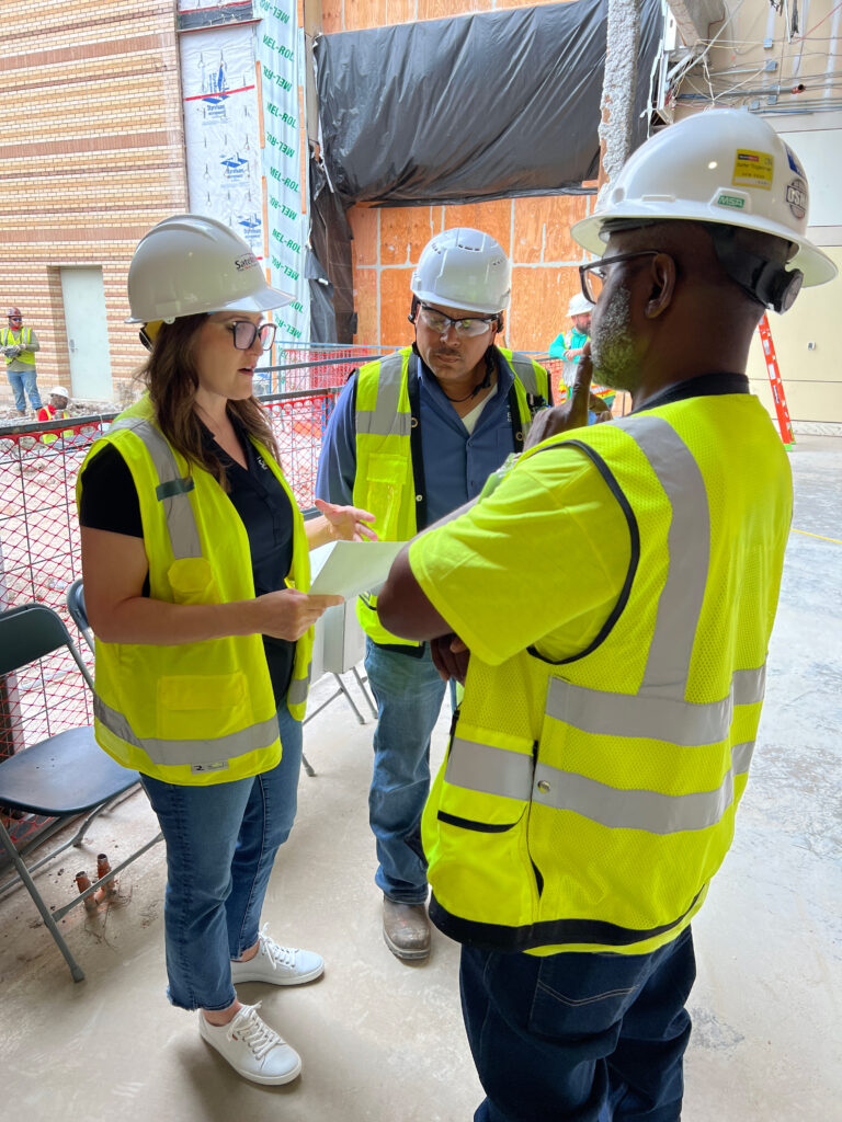 A female construction worker in a white hard hat and yellow safety vest holding a piece of paper and leading 2 male colleagues.