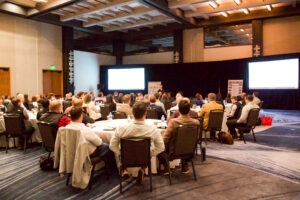 team members sitting at round tables in a large meeting room with a speaker standing in the front 