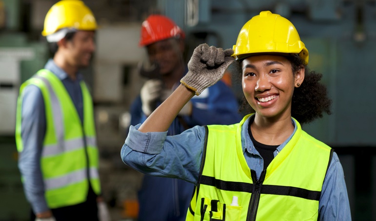 A smiling female construction worker wearing a yellow hard hat and safety vest is adjusting her hat.