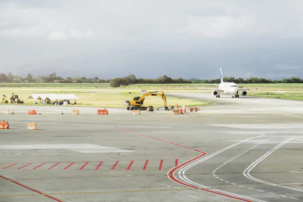 Construction crews and equipment work near an active runway as a commercial airplane taxis on a cloudy day.