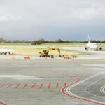 Construction crews and equipment work near an active runway as a commercial airplane taxis on a cloudy day.
