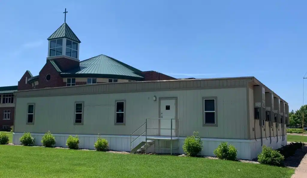 Four connected portable classrooms sit in front of a church, with a steeple in the background.