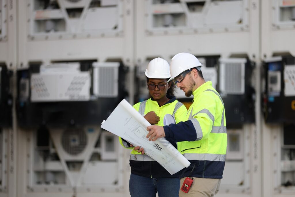 Two construction professionals reviewing blueprints in front of large equipment panels
at an industrial job site.