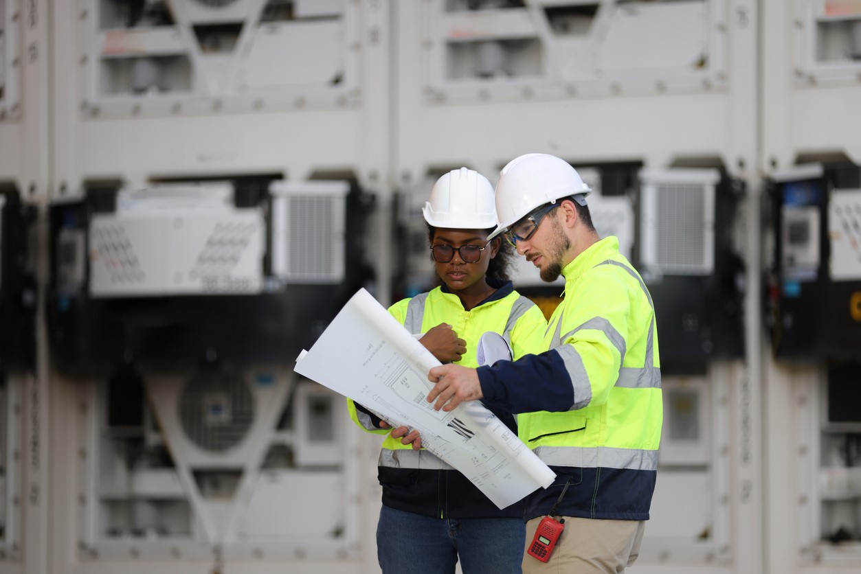 Two construction professionals reviewing blueprints in front of large equipment panels at an industrial job site.