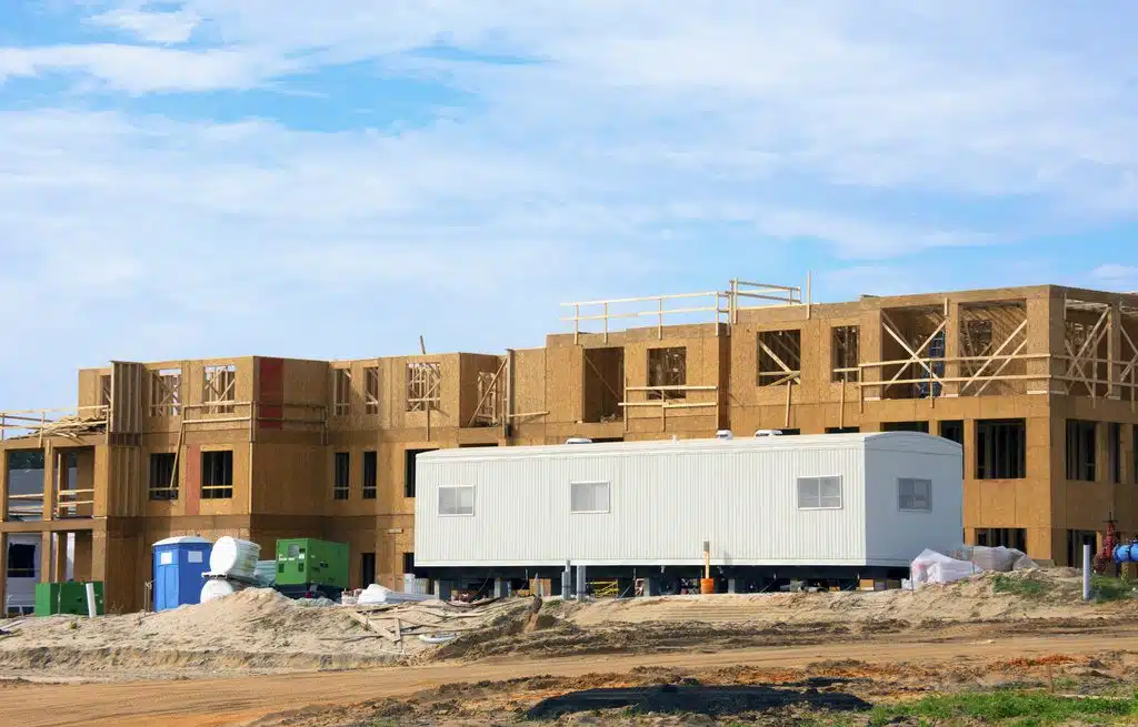 Large construction site half constructed wooden building with mobile job location office trailer in front area for contractors, work managers and supervisors for paperwork and worker interviews.
